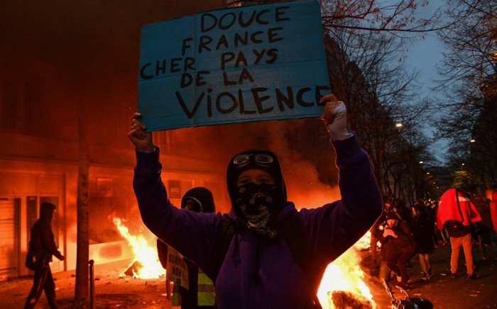 A protester holds a sign reading "Sweet France. Dear country of violence" in front of a fire during a demonstration in Paris on 5 December, 2020. Picture: AFP. 