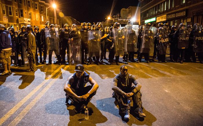Two protesters sit on the ground in front of riot police minutes before a mandatory, city-wide curfew of 10 pm near the CVS pharmacy that was set on fire during rioting after the funeral of Freddie Gray, on 28 April, 2015 in Baltimore, Maryland. Picture: AFP.